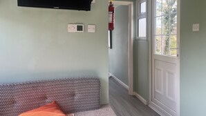 Quarto com televisão de tela plana na parede, termostato, extintor de incêndio e porta que dá para uma varanda com vista para árvores.