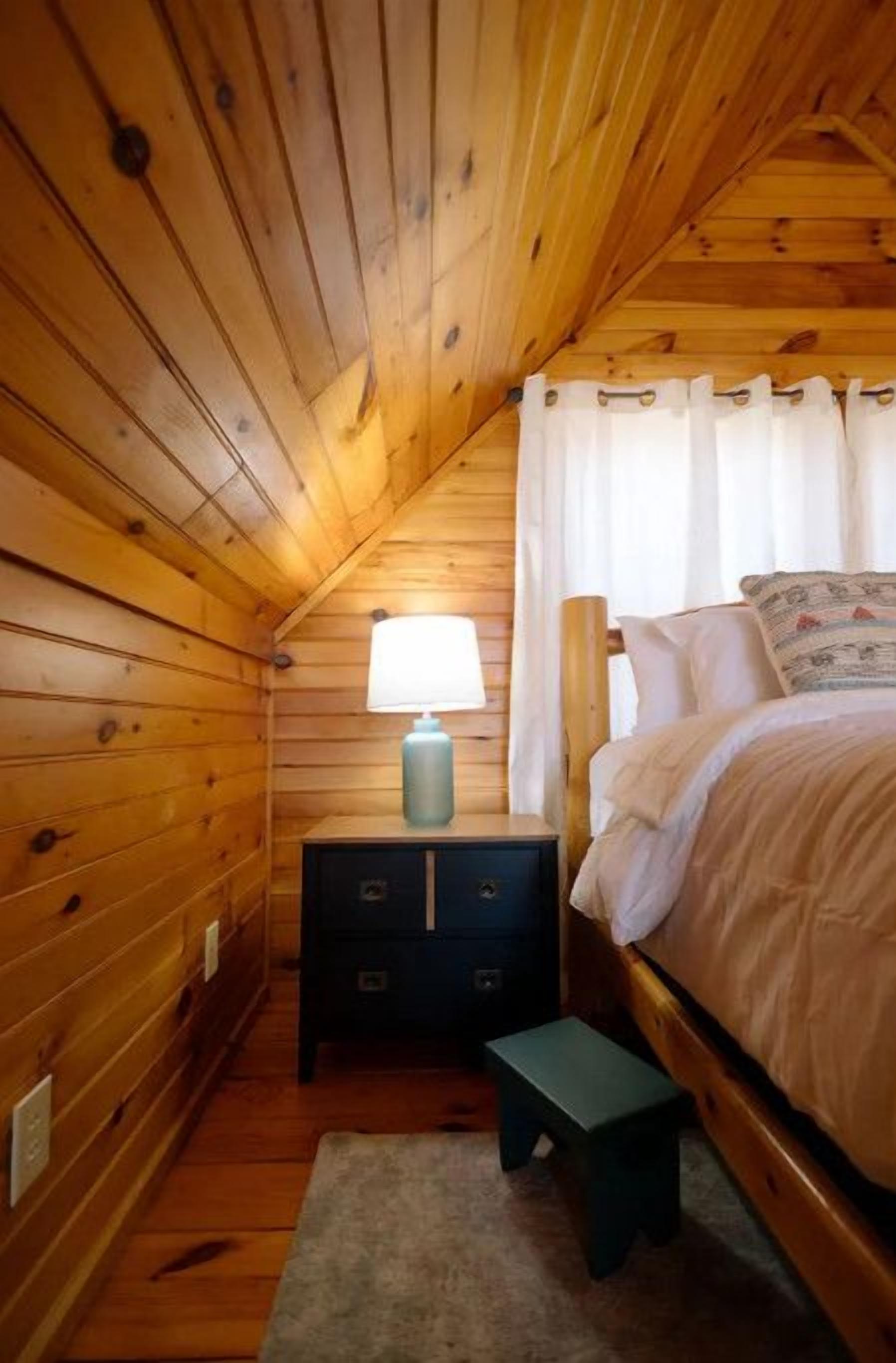 A wooden cabin bedroom with a bed, nightstand, lamp, and a small stool.