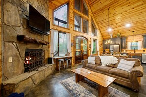 Interior acogedor de una cabaña con chimenea de piedra, techo de madera, sala de estar con sofá y mesa de centro, y zona de cocina.