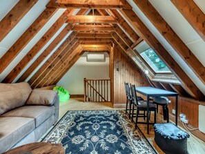 An attic room with a sloped ceiling, wooden beams, a sofa, a dining table with chairs, a rug, and a skylight.