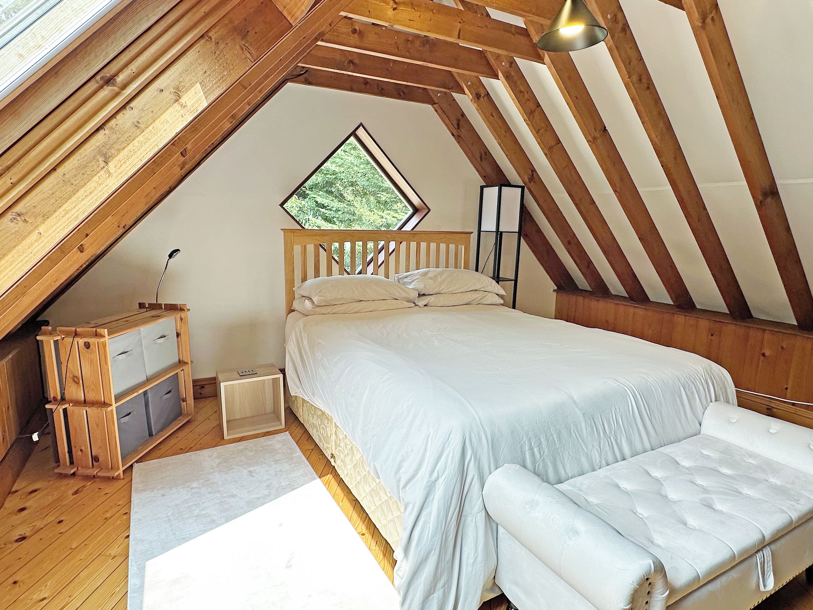 A bedroom with a sloped ceiling, a bed with white bedding, a bedside table, a lamp, and a window with a view of greenery.