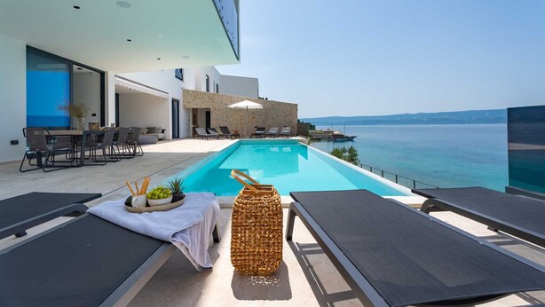 A modern outdoor pool area with lounge chairs, a dining setup, and a view of the sea. - Villa Sea Breeze, 5 bedroom Luxury Villa near the sea, private heated pool, Omiš Brzet (Omiš)