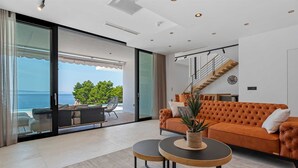 A modern living room with a large sliding glass door leading to a balcony with a view of the sea. There is a sofa, a coffee table, and a staircase to the upper floor. - Villa Sea Breeze, 5 bedroom Luxury Villa near the sea, private heated pool, Omiš Brzet (Omiš)