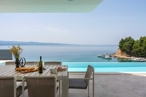 Una mesa de comedor con una botella de vino, una copa y un pan, con vista a un mar azul cristalino con barcos y un acantilado.