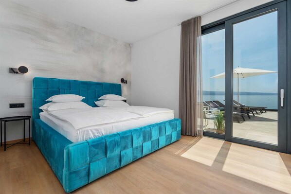 Un dormitorio moderno con una cama grande, vista al mar a través de una puerta corrediza de vidrio y una pequeña mesita de noche.