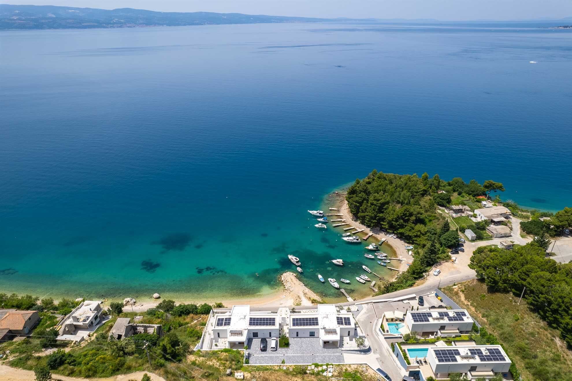 An aerial view of a coastal area with a beach, boats, and modern buildings with solar panels.