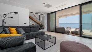 Een moderne woonkamer met een grijze bank, een salontafel en uitzicht op zee door grote glazen deuren.