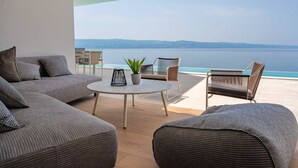Een moderne woonkamer met een grijze bank, twee fauteuils, een ronde tafel en een potplant. De kamer heeft een groot raam met uitzicht op een waterpartij en heuvels in de verte.