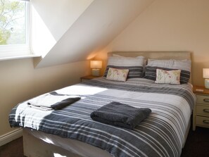 Ein Schlafzimmer mit schrägen Wänden, einem Bett mit gestreifter Bettwäsche, zwei Nachttischlampen und einem Fenster mit Blick auf Bäume.