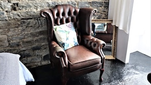 A brown leather armchair with a floral pillow against a stone wall, a mirror with a gold frame, and a white curtain partially visible.
