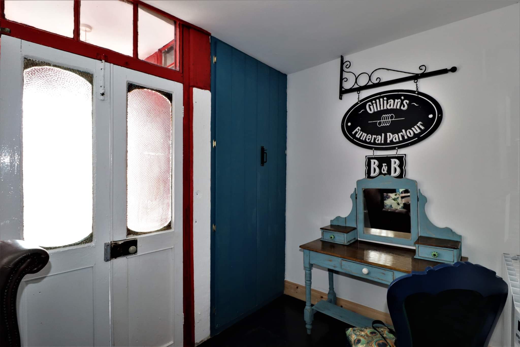A room with a white door, a blue door, a blue wall, a small table with a mirror, and a sign above the table.