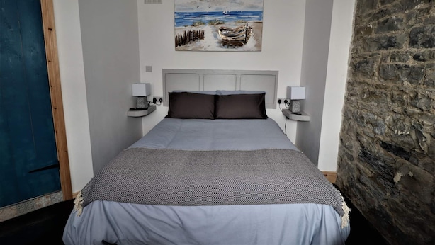 A bedroom with a bed, a painting of a beach scene, a stone wall, and a blue door.