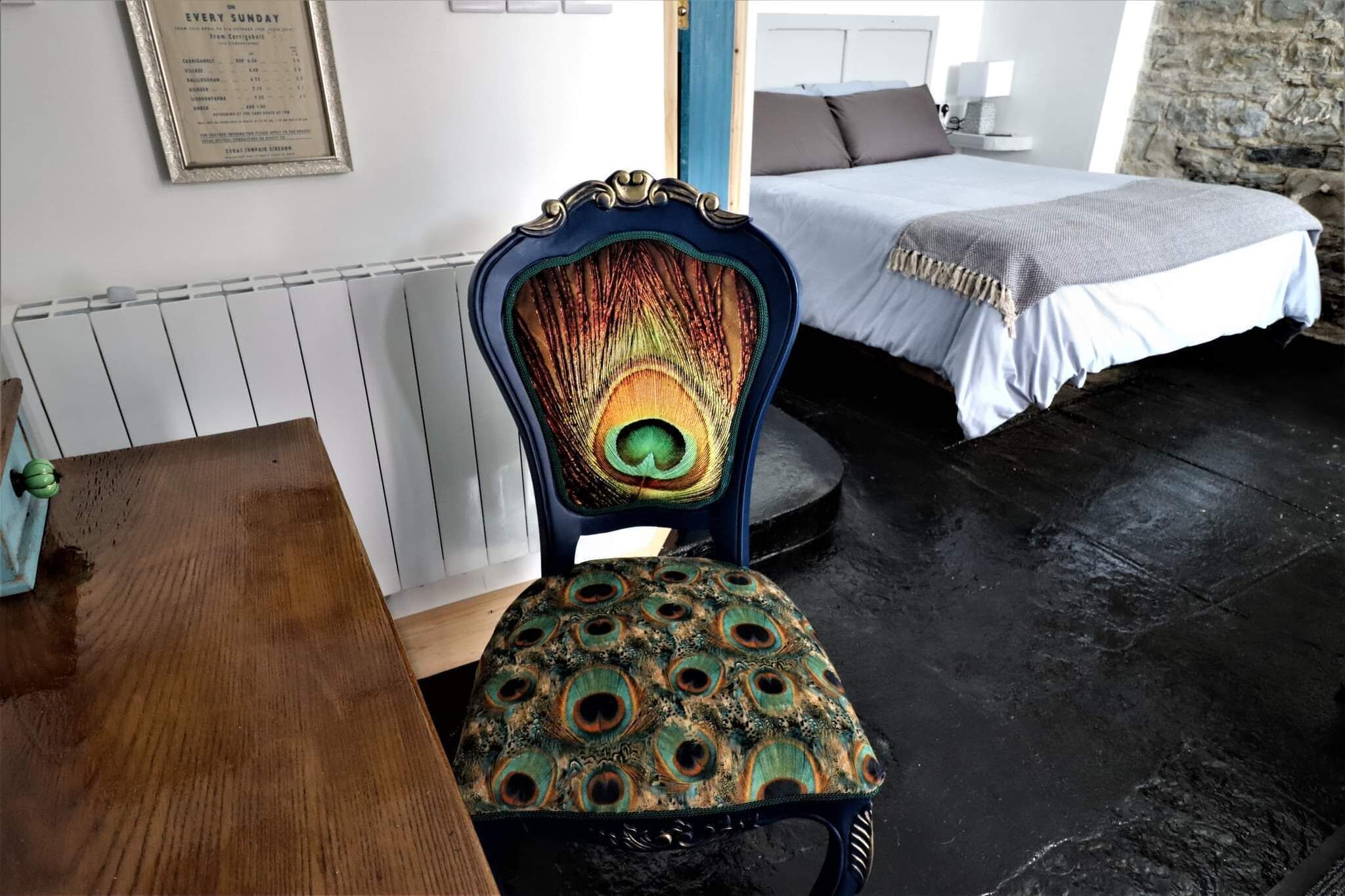 A room with a bed, a chair with a peacock design, and a wooden table.