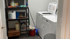 A laundry area with a washing machine, a shelf with cleaning supplies, and a trash bin. - Cozy Lake View Condo (Storm Lake)