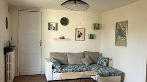 Ein Wohnzimmer mit einer Eckcouch, zwei kleinen runden Tischen, einem Heizkörper, einer Tür und Wanddekorationen, darunter eine Uhr und gerahmte Kunstwerke.