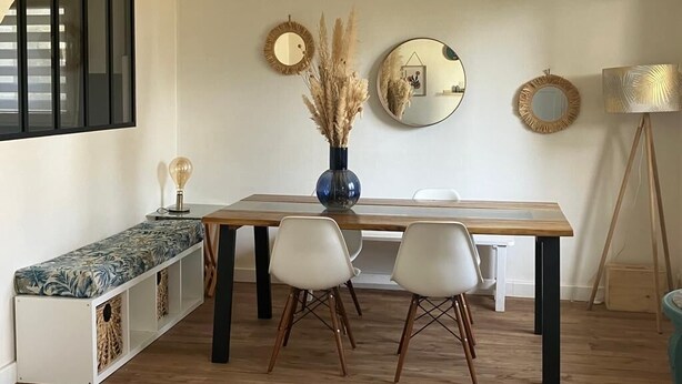 Ein Essbereich mit einem Holztisch, zwei weißen Stühlen, einer Vase mit Trockenpflanzen und drei Wandspiegeln.