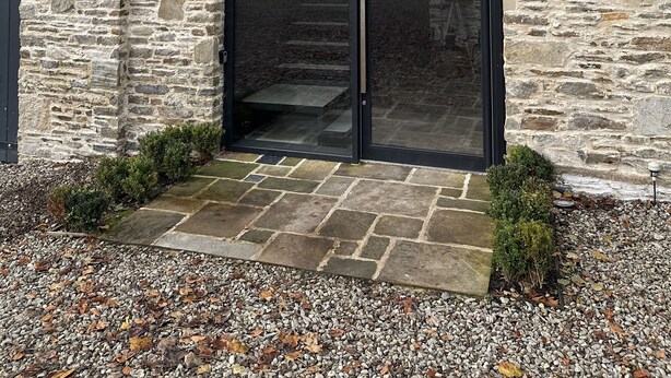 Ein Steinhaus mit einer großen Glasdoor und einem kleinen Gartenbereich mit Kies und Pflanzen.
