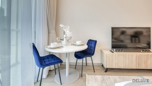 Ein modernes Wohnzimmer mit einem runden Esstisch, zwei blauen Stühlen, einer weißen Vase mit Blumen, einem Flachbildfernseher auf einem Holzständer und einem großen Fenster mit transparenten Vorhängen.