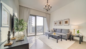 A modern living room with a grey sofa, a coffee table, and a TV on a wooden cabinet. There is a large window with curtains, a chandelier, and framed pictures on the wall. - Graceful 1BR at Park Heights Dubai Hills MBR City by Deluxe Holiday Homes (Dubai)