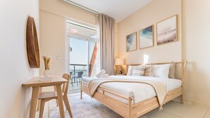 Quarto de hotel com cama, escrivaninha e cadeira. Há uma janela com cortinas, uma porta e uma varanda.