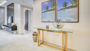 Uma sala de estar moderna com uma mesa de centro de vidro, uma pintura grande com uma paisagem praiana e um vaso em uma mesinha lateral.