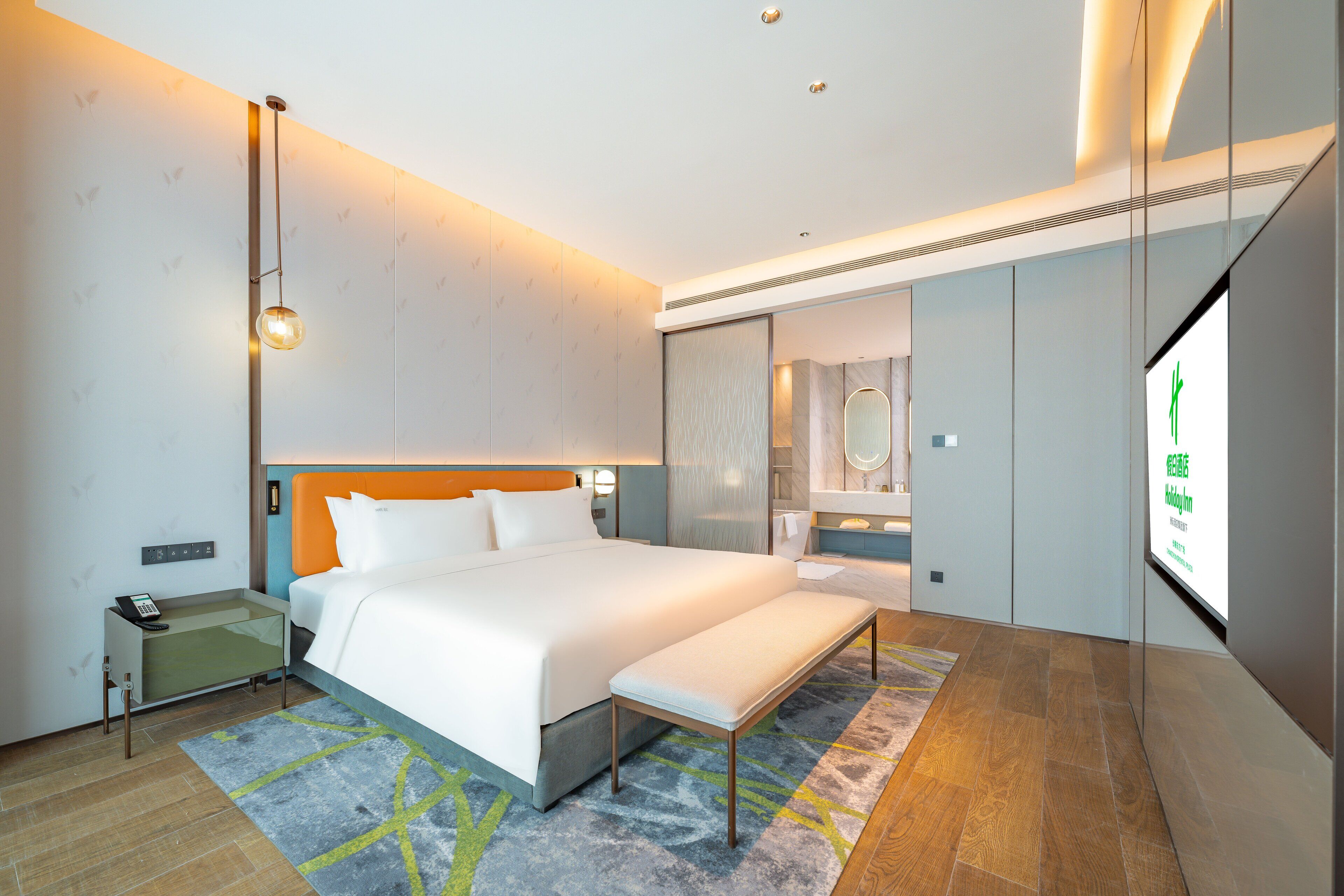 A modern hotel room with a large bed, bedside table, bench, and a TV screen displaying text.