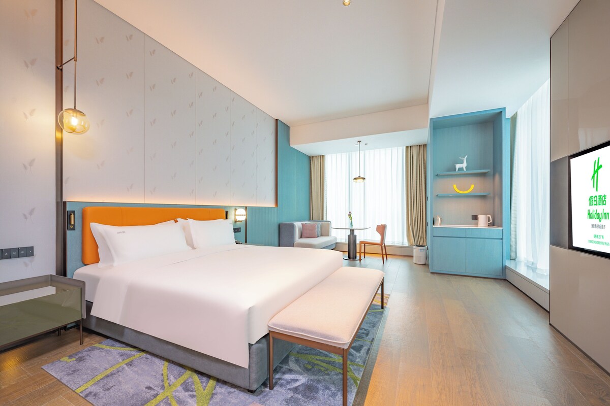 Une chambre d’hôtel moderne dotée d’un grand lit, d’une banquette, d’un bureau avec une chaise, d’une télévision et d’une petite kitchenette.