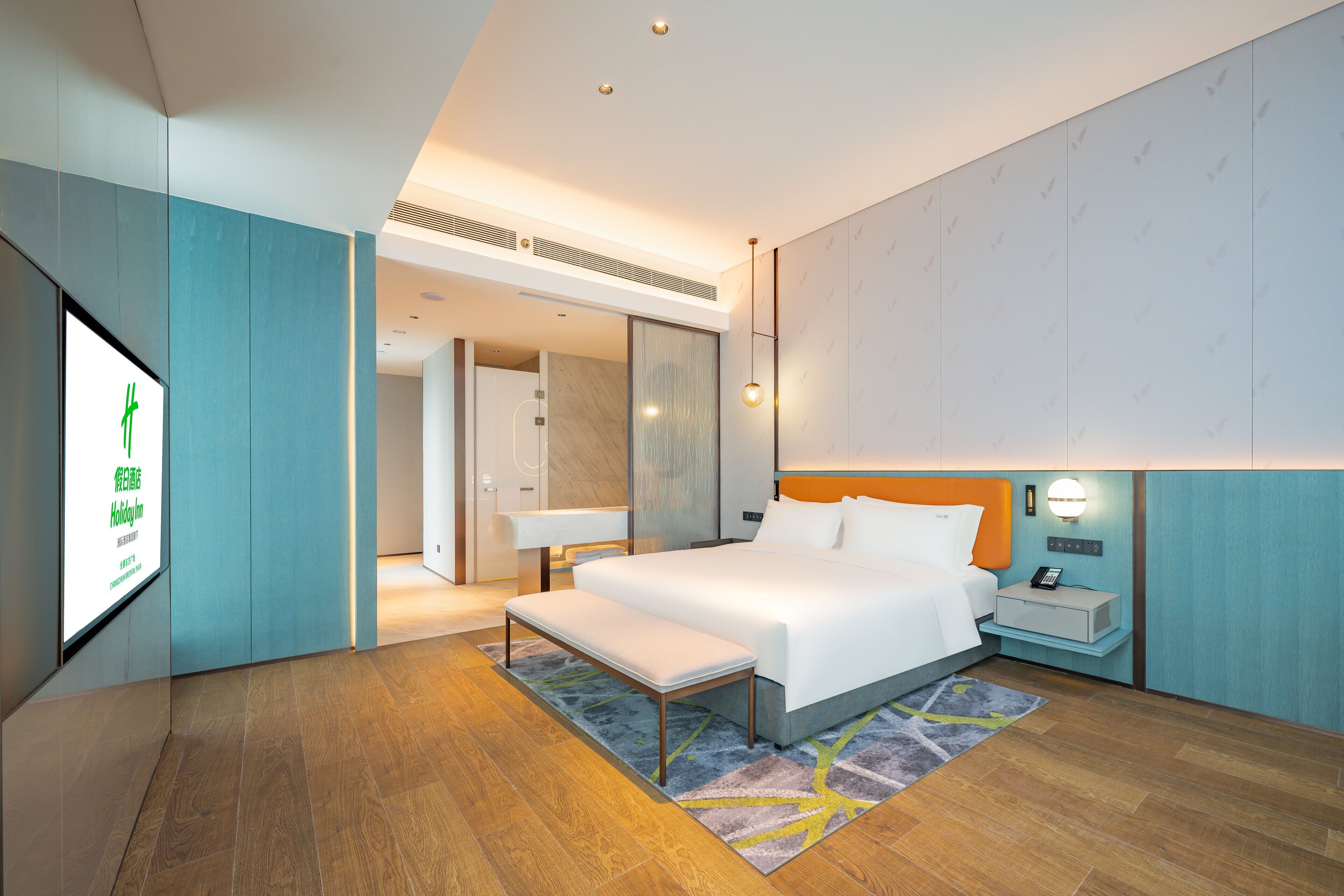 Ein modernes Hotelzimmer mit einem großen Bett, einer Bank, einem Nachttisch und einem Fernseher, der eine Speisekarte zeigt.