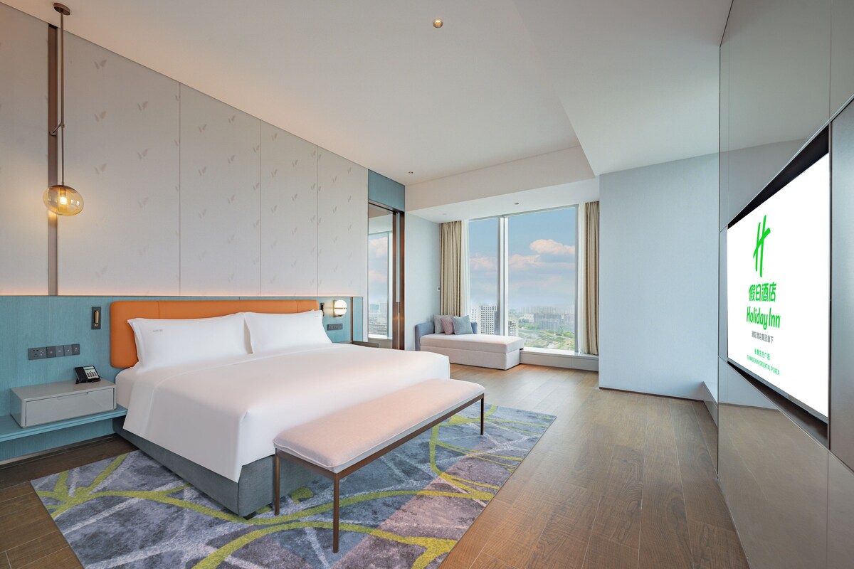 Une chambre d’hôtel moderne dotée d’un grand lit, d’une table de chevet, d’une petite table, d’une grande fenêtre donnant sur la ville et d’un téléviseur fixé au mur qui affiche des informations sur l’hôtel.