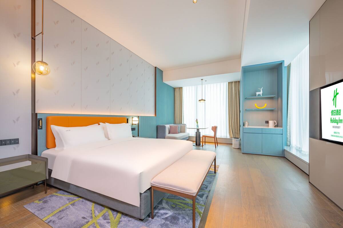 Een moderne hotelkamer met een groot bed, een bankje, een bureau met stoel, een televisie en een kleine kitchenette.