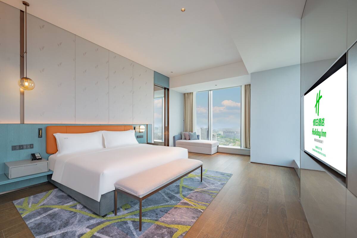 Een moderne hotelkamer met een groot bed, een nachtkastje, een groot raam met uitzicht op de stad en een aan de muur bevestigde televisie waarop hotelinformatie wordt getoond.