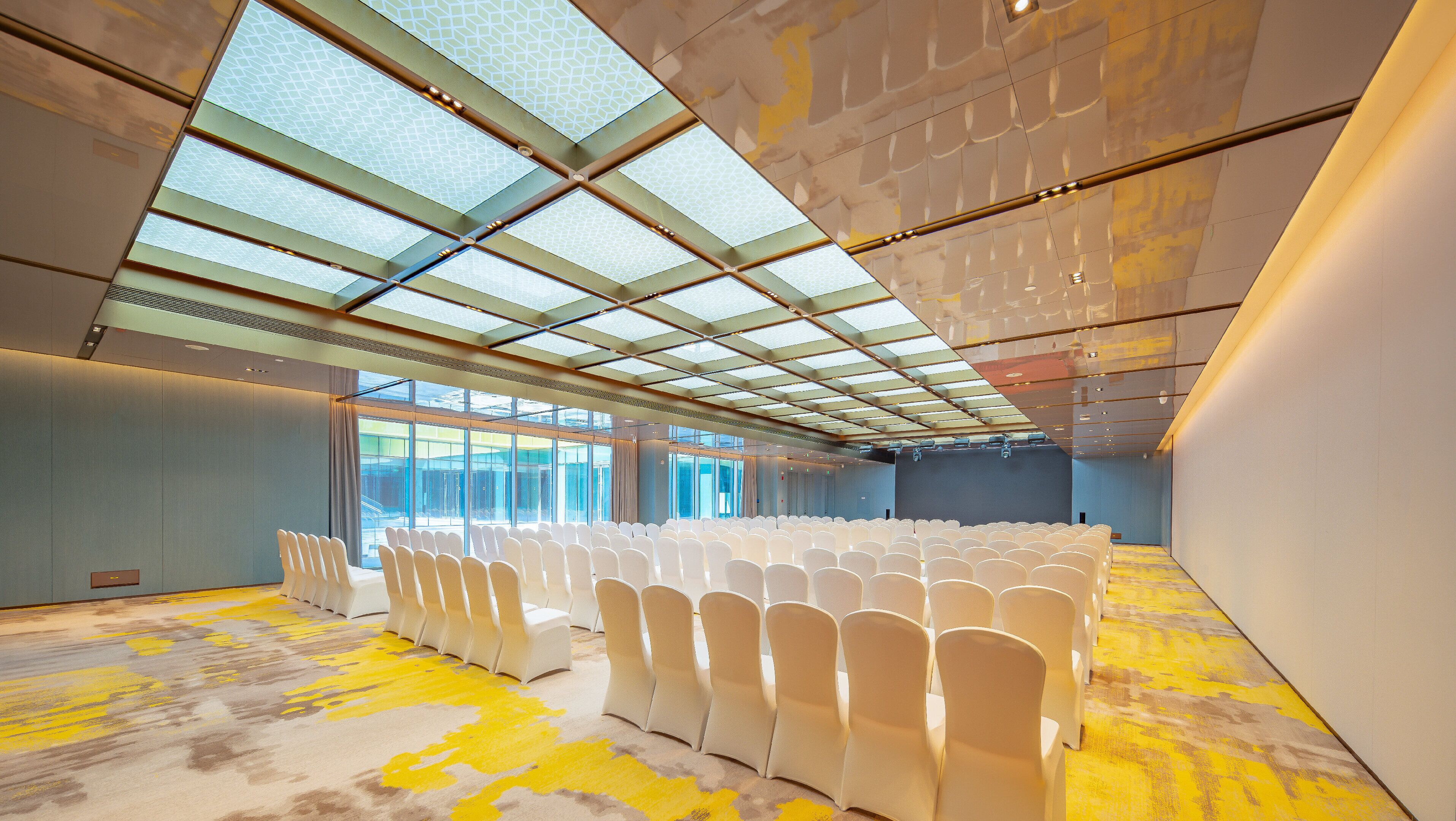 Un salón de conferencias amplio con filas de sillas blancas, un techo alto con tragaluces y una alfombra con diseño amarillo.