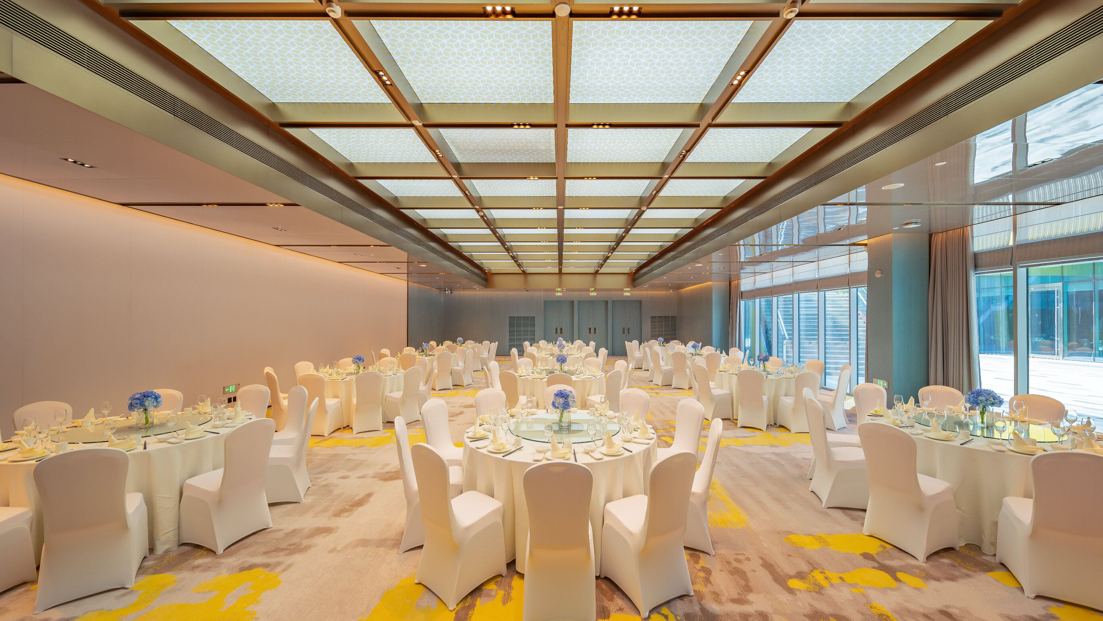 Een ruime congrescentrum met ronde tafels, witte stoelen en een hoog plafond met dakramen.