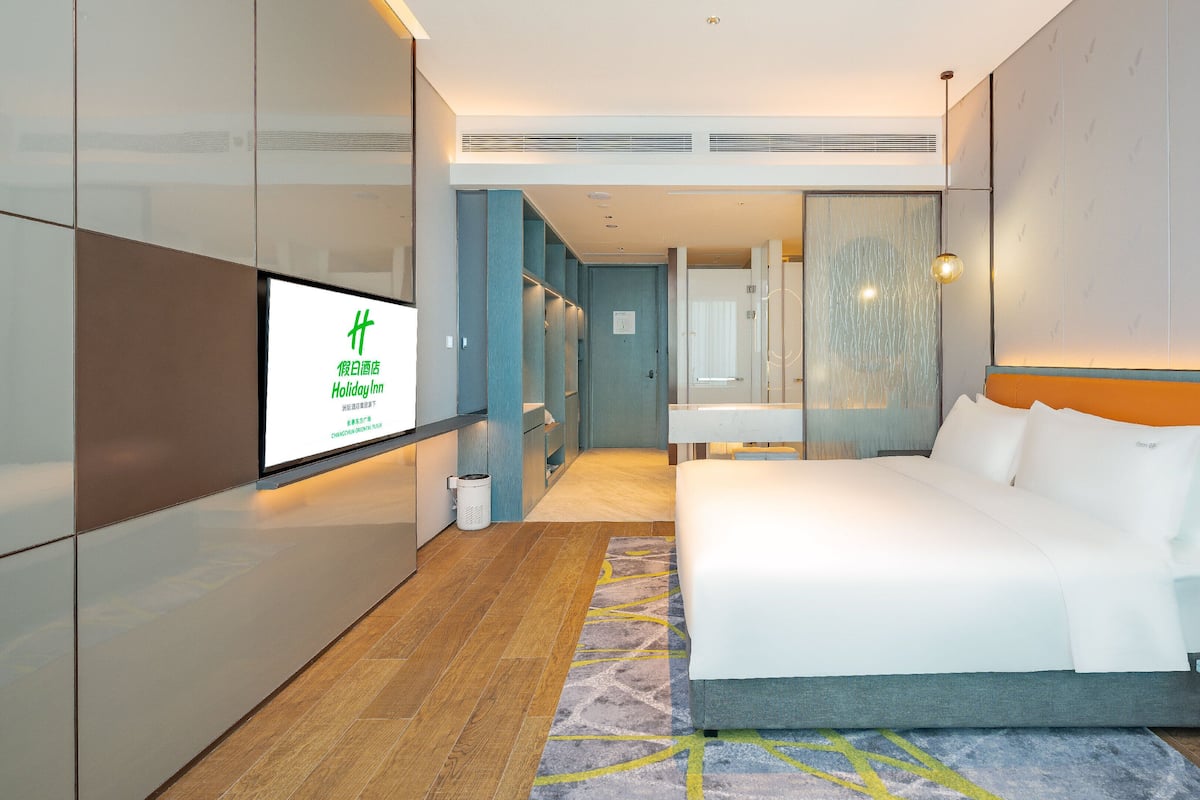 Een moderne hotelkamer met een groot bed, een televisie waarop een hotelsignaal staat, een badkamer met spiegel, en een gang die naar een andere kamer leidt.