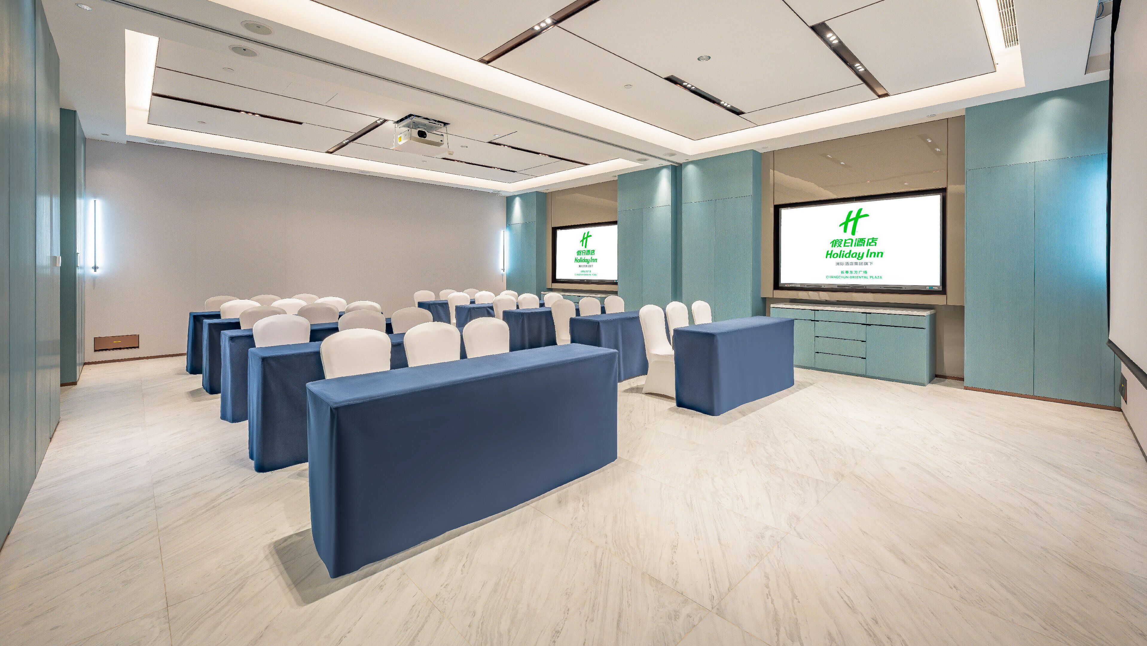 A conference room with rows of tables and chairs, a projector, and a screen displaying text.