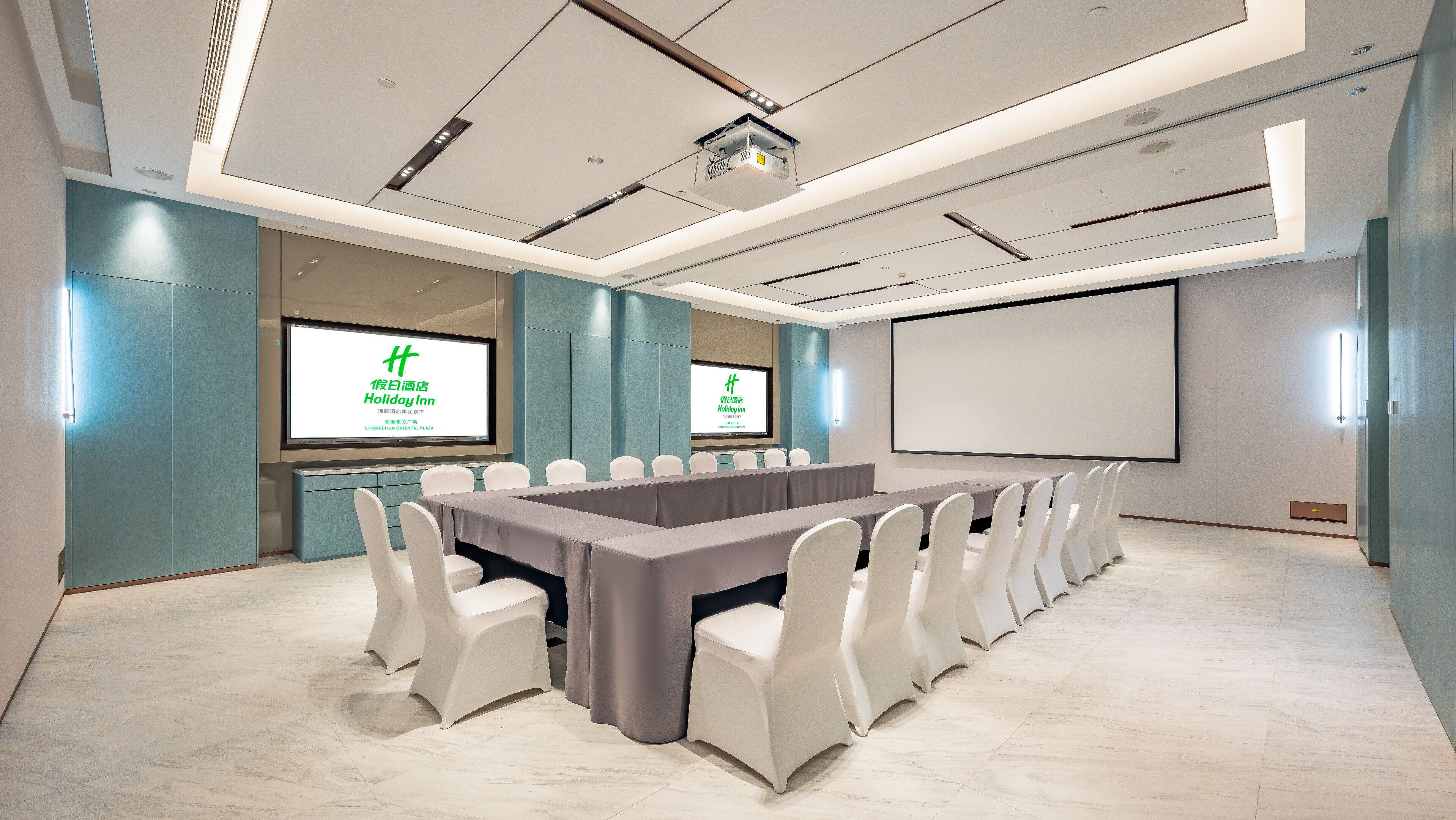 A conference room with a long table, white chairs, a projector screen, and a large screen displaying a hotel name.
