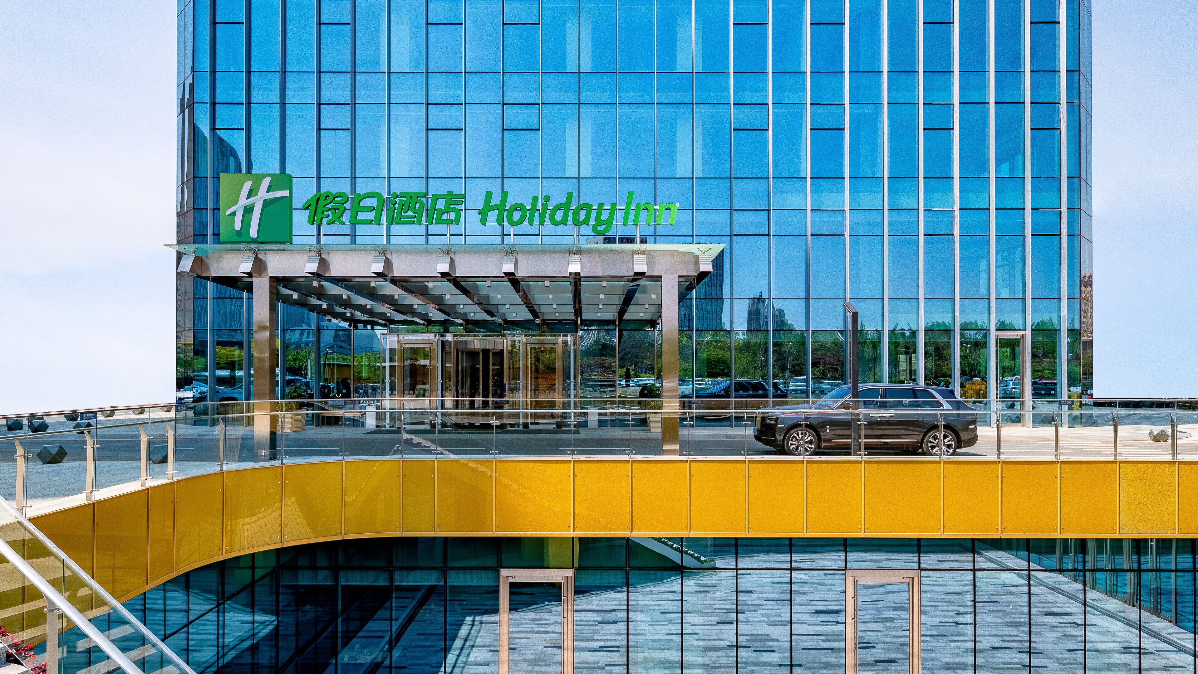 A Holiday Inn hotel with a modern glass facade, a covered entrance, and a yellow pedestrian bridge.