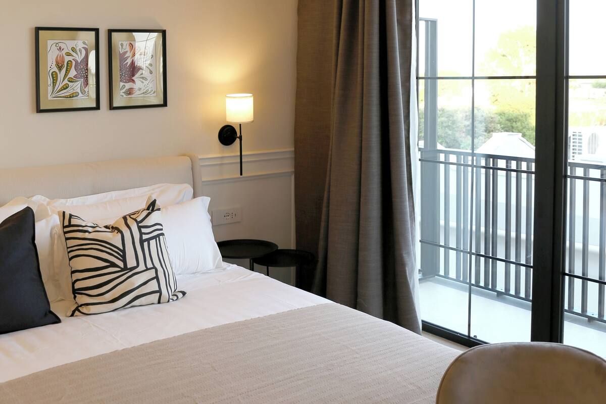 A hotel room with a bed, two pillows, a bedside table with a lamp, a chair, and a large window with a view of the outside.