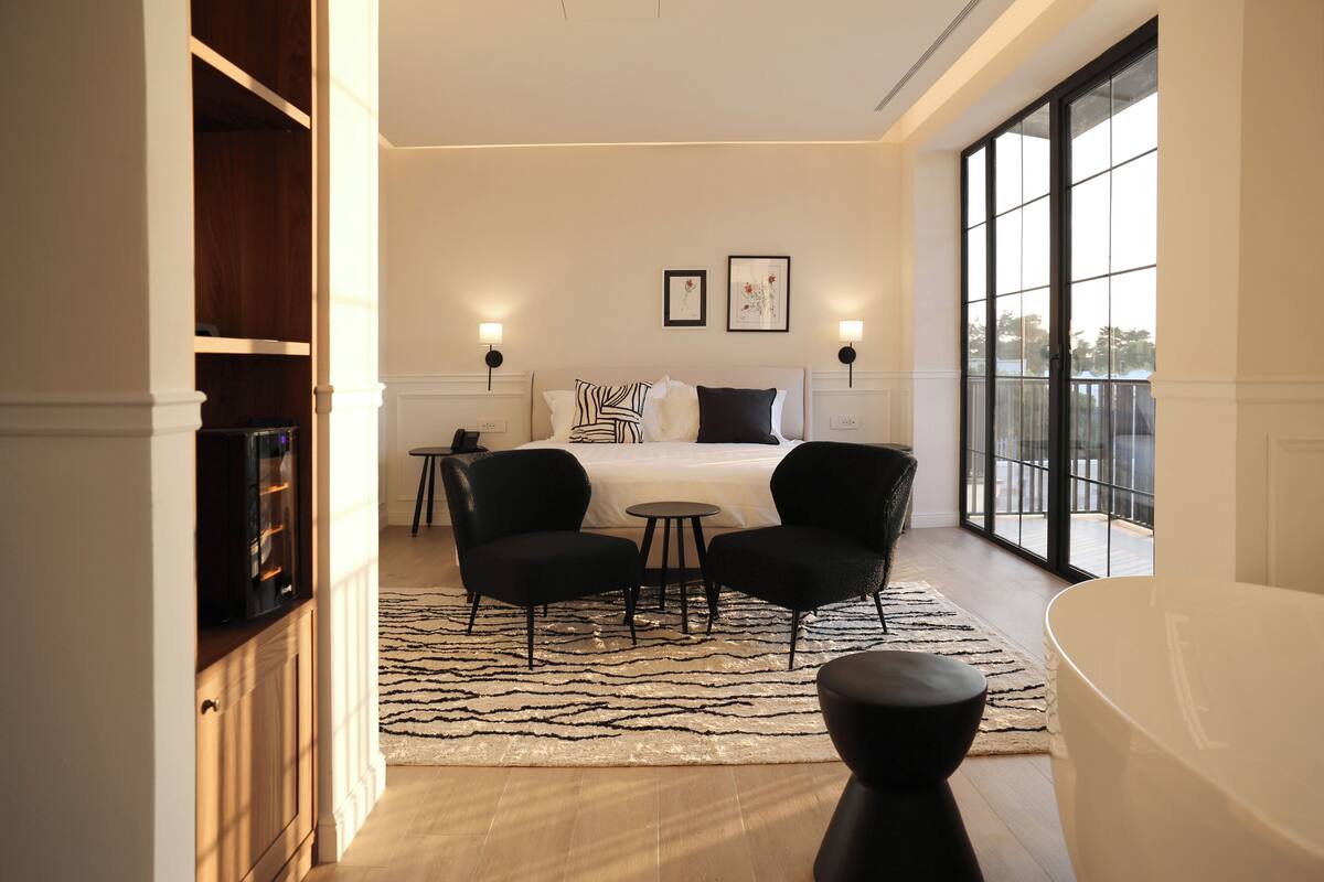 A bedroom with a bed, two chairs, a small table, a large window, and a bathtub.