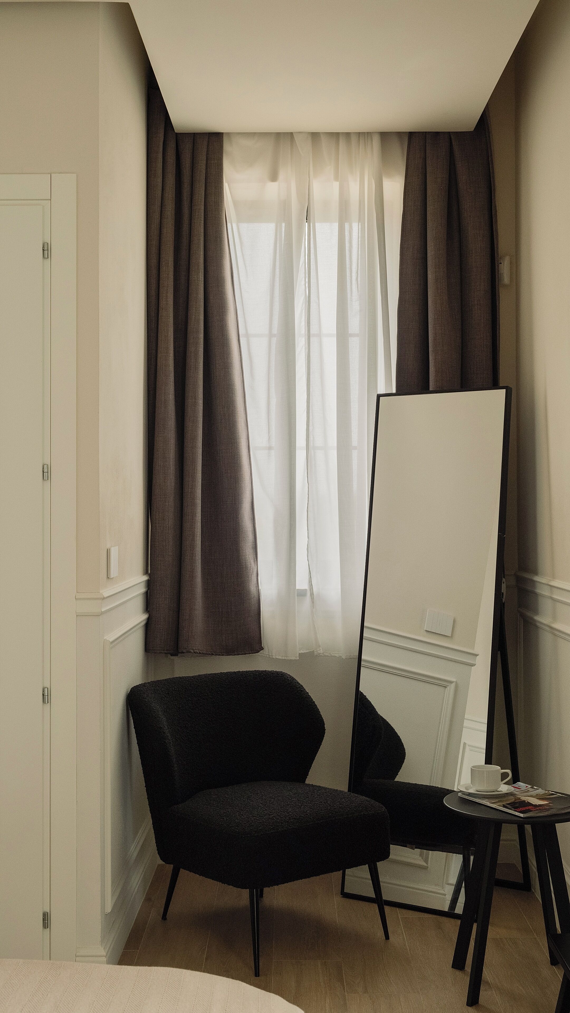 A room with a large mirror, a black chair, a small table with a cup and a book, and a window with curtains.