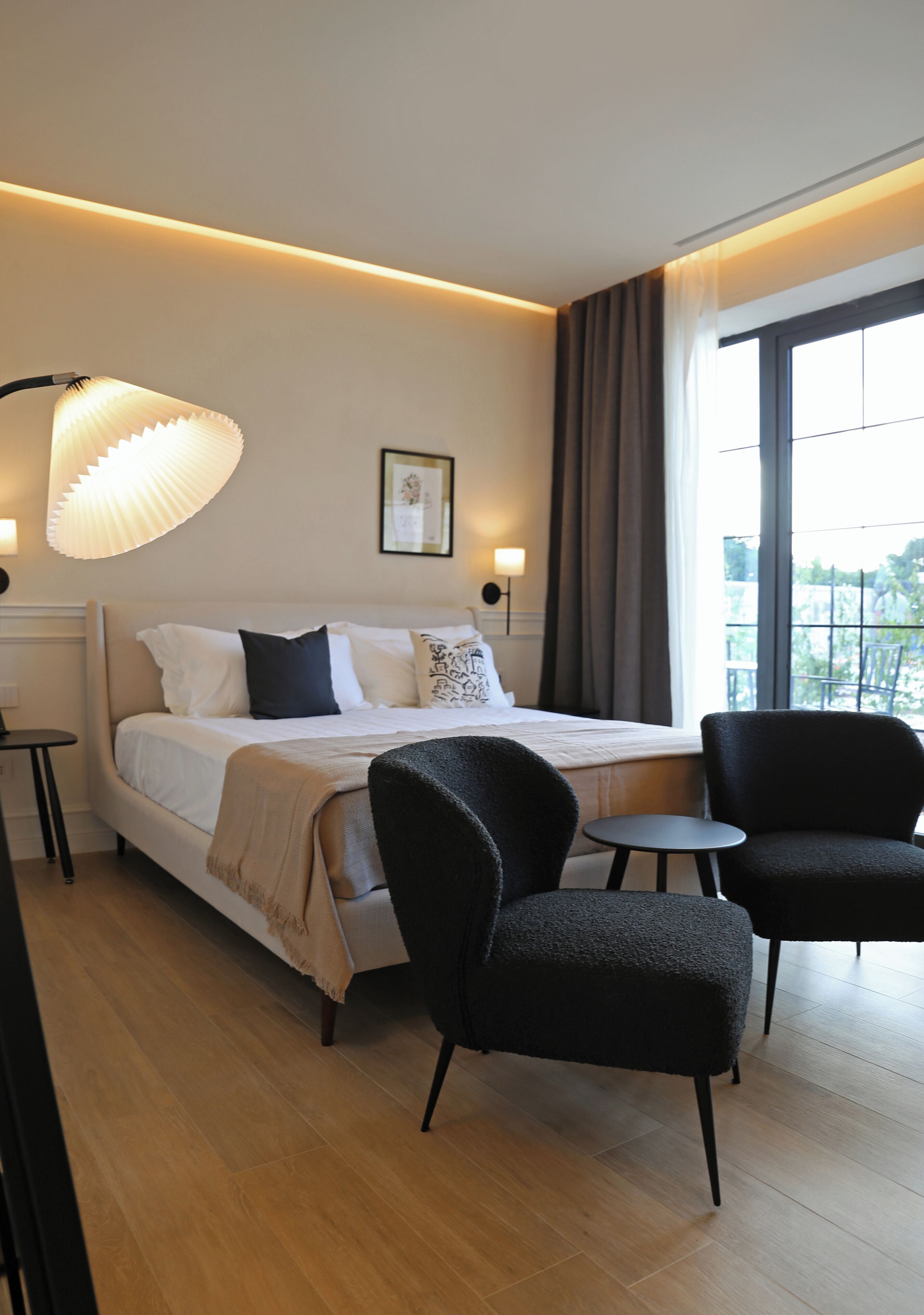 Une chambre d’hôtel moderne, dotée d’un grand lit, de deux fauteuils, d’une petite table et d’une grande fenêtre offrant une vue dégagée.