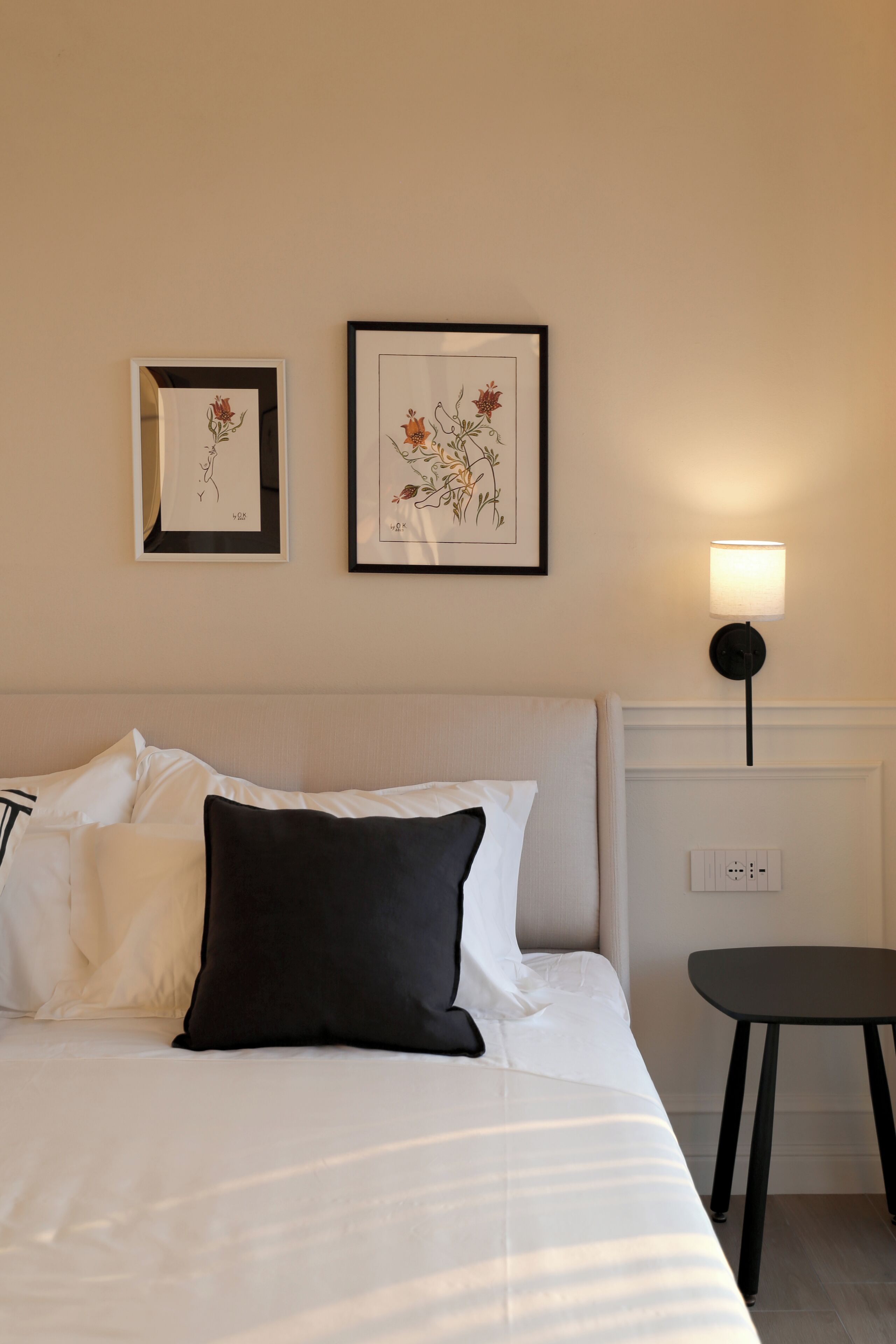 A neatly made bed with white and black pillows, a small black table beside it, and two framed floral artworks on the wall.