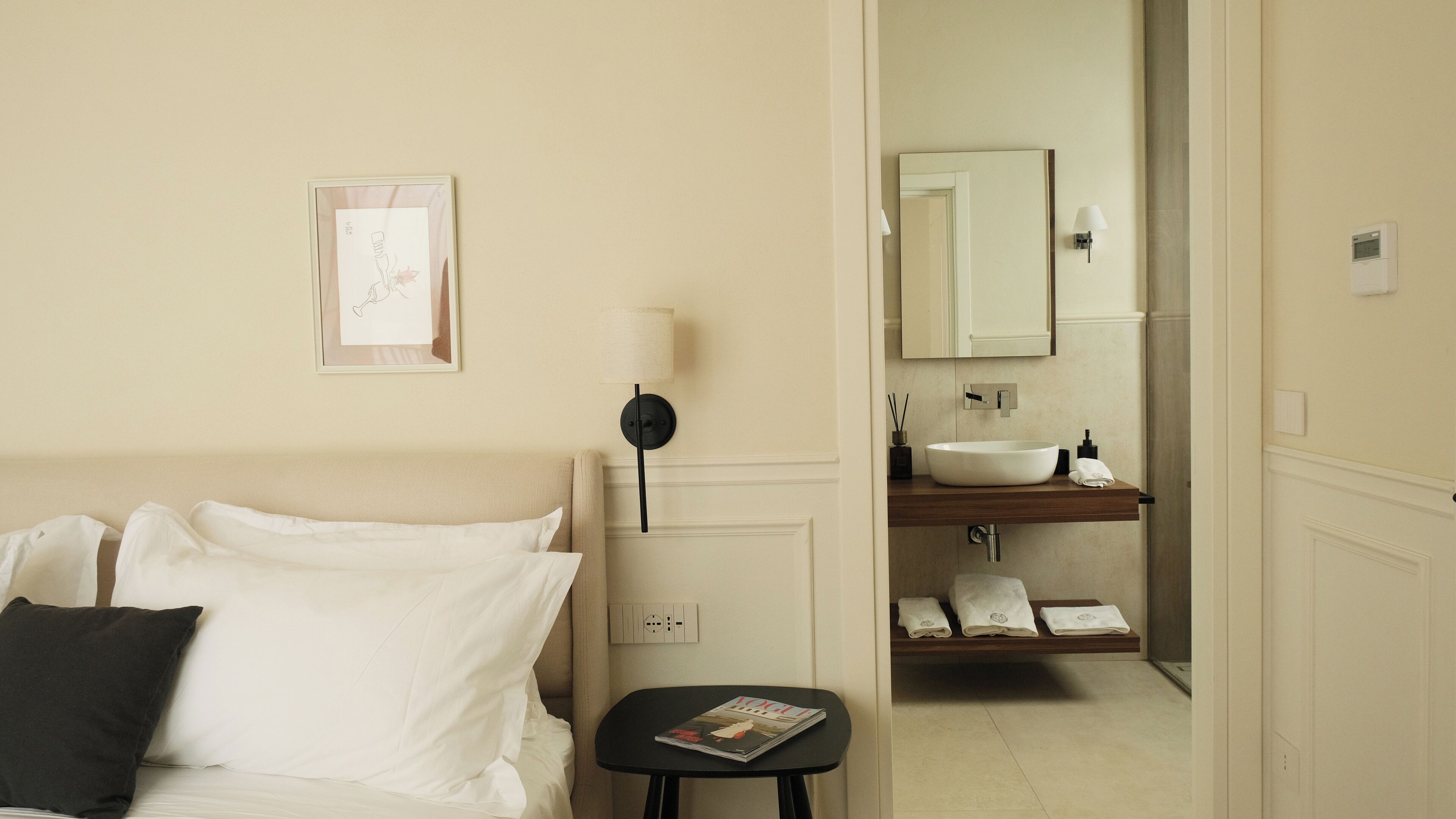 Quarto de hotel com uma cama, mesa de cabeceira com uma revista, quadro pendurado na parede e banheiro visível através de uma porta aberta.