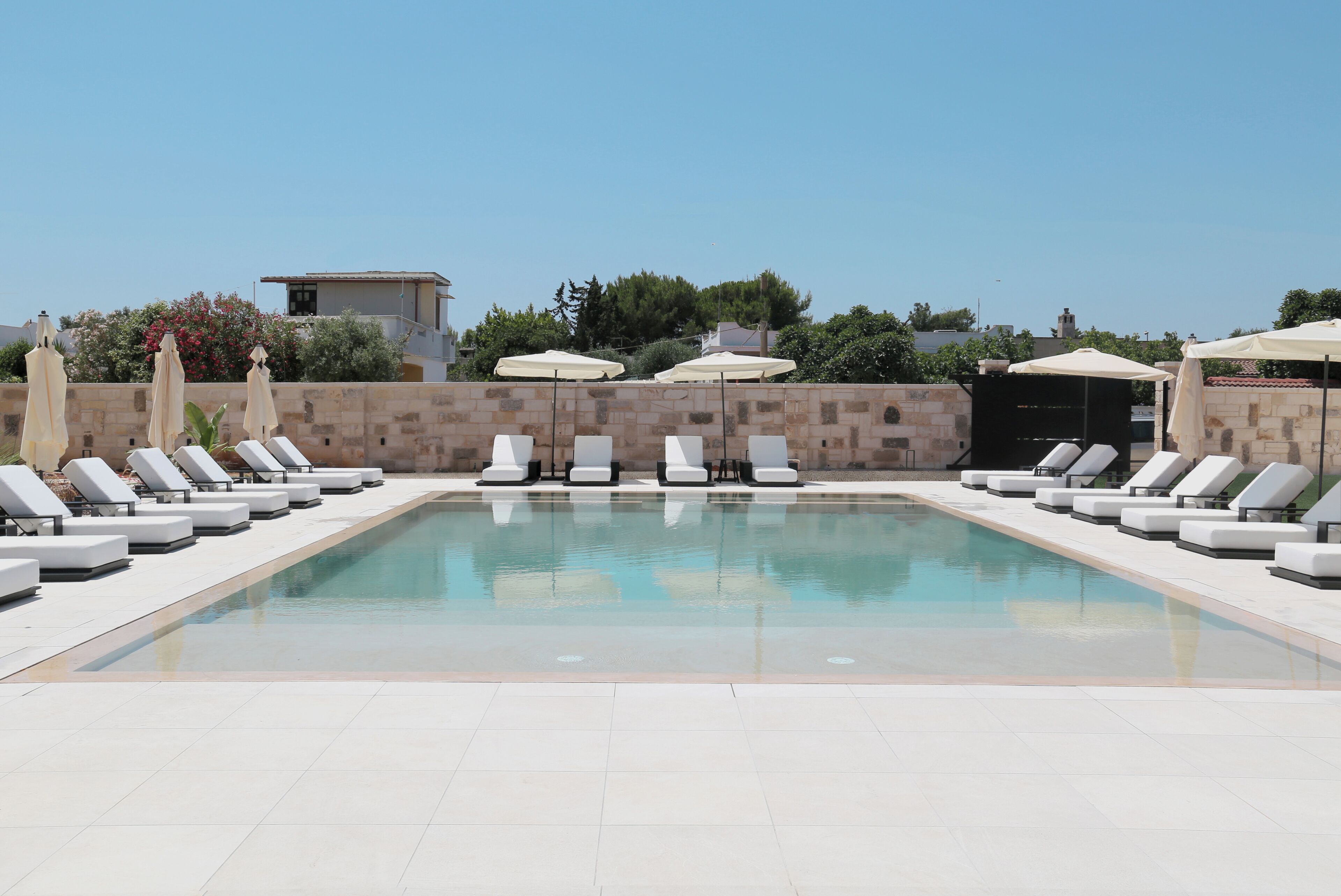 A rectangular swimming pool with white lounge chairs and umbrellas on either side, surrounded by stone walls and trees.