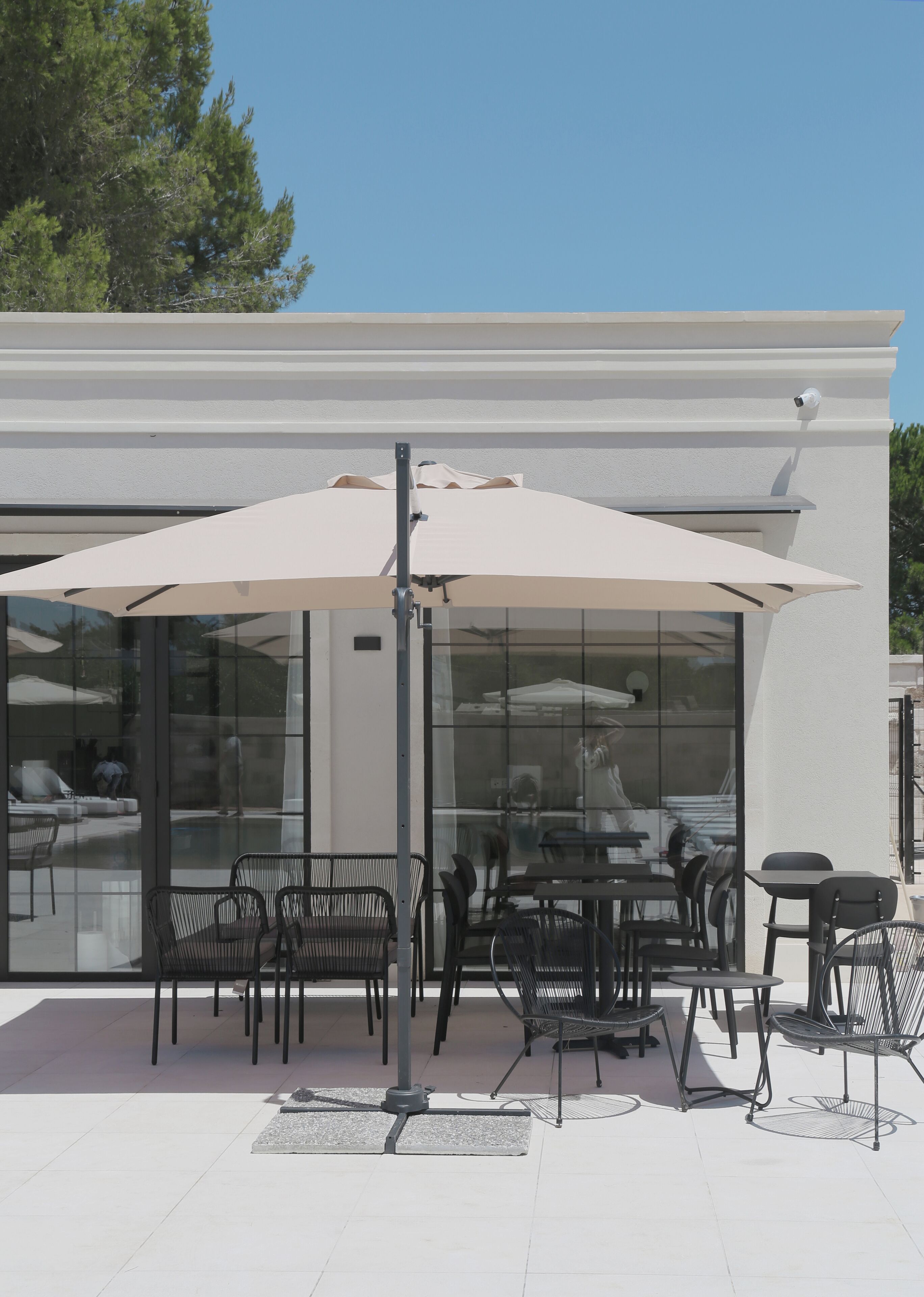 An outdoor patio with tables and chairs, a large umbrella, and a modern building in the background.
