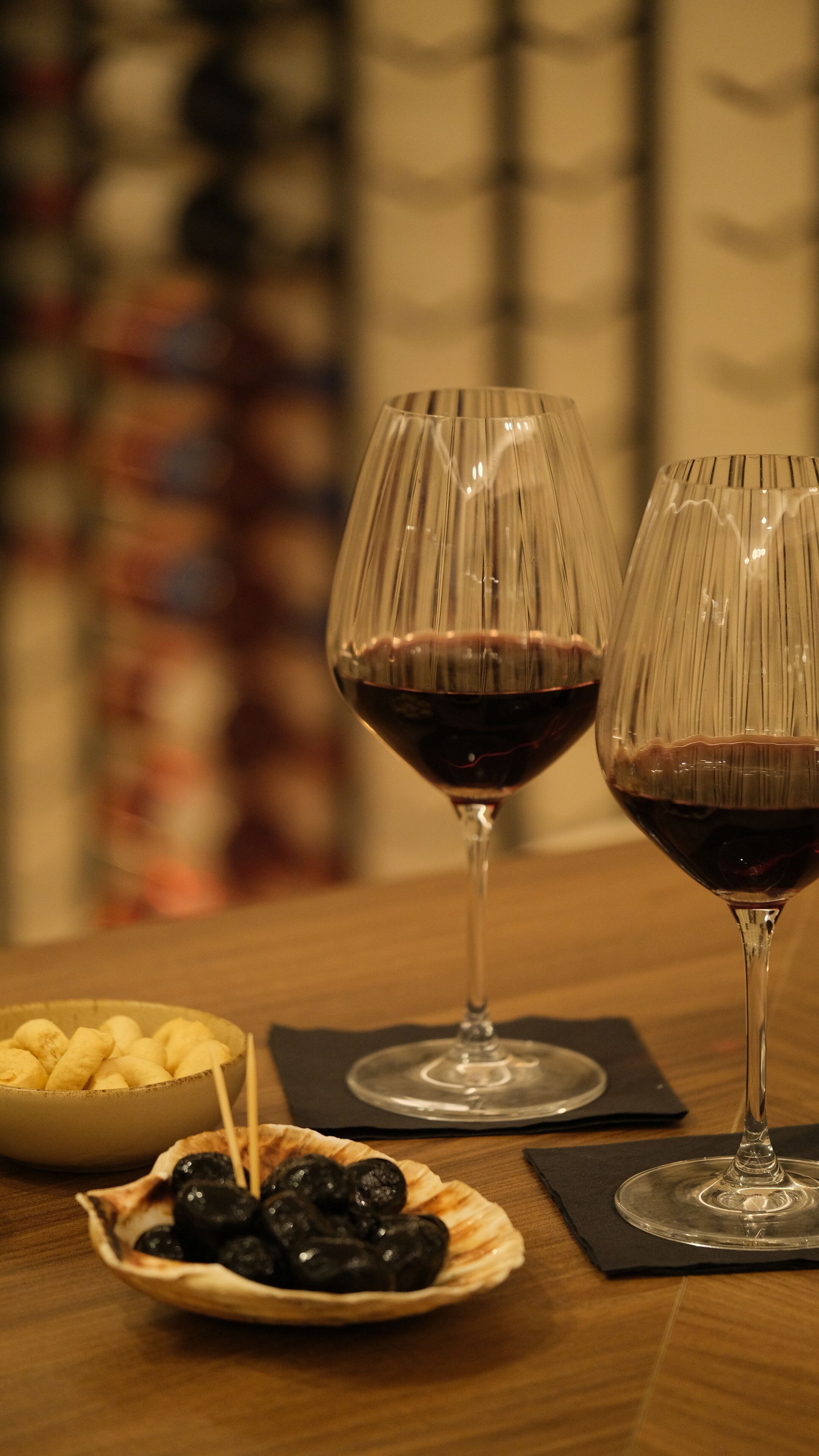 Two wine glasses on a table with a bowl of olives and a bowl of cheese sticks.