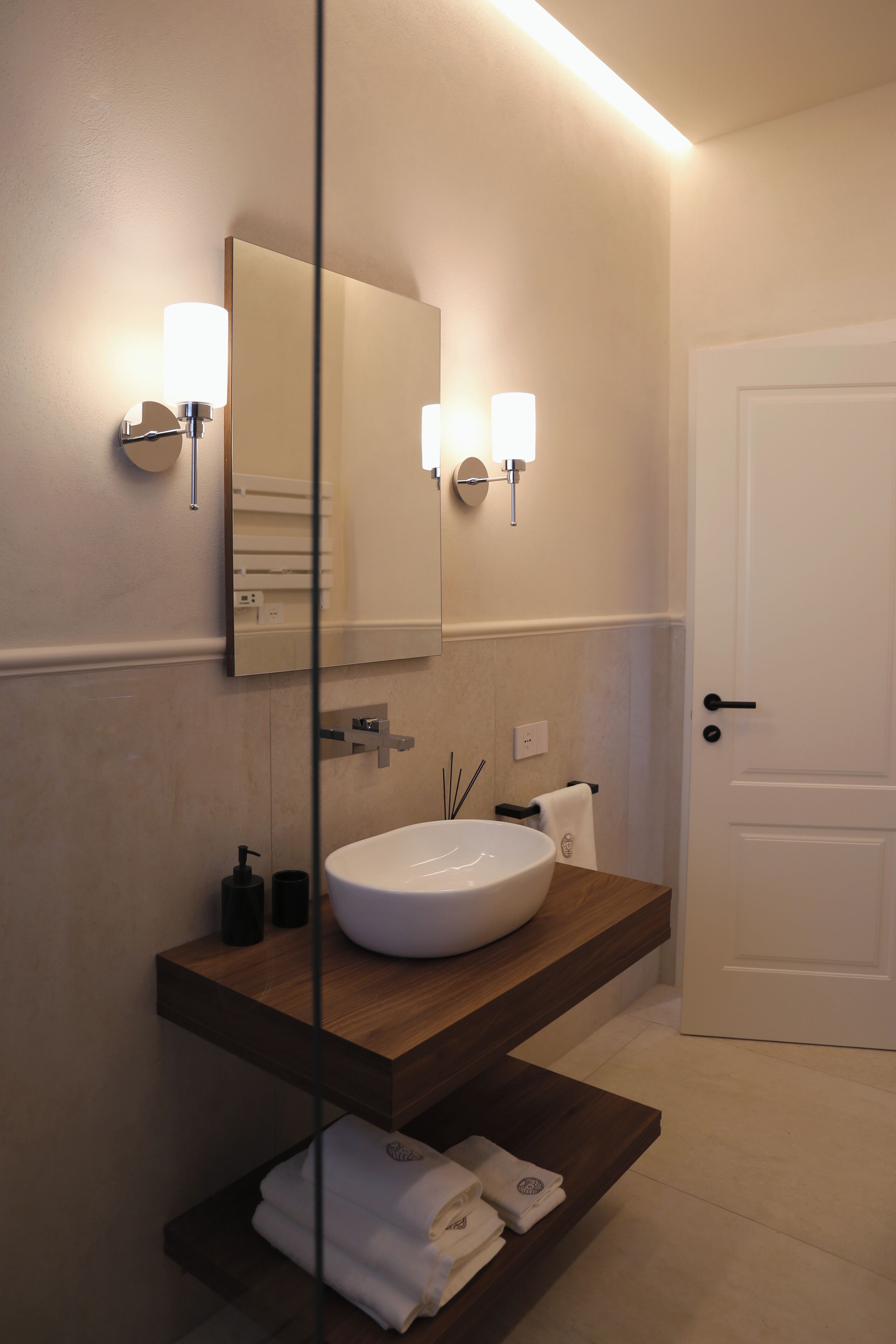 A modern bathroom with a large mirror, wall-mounted lights, a wooden vanity, and a white door.