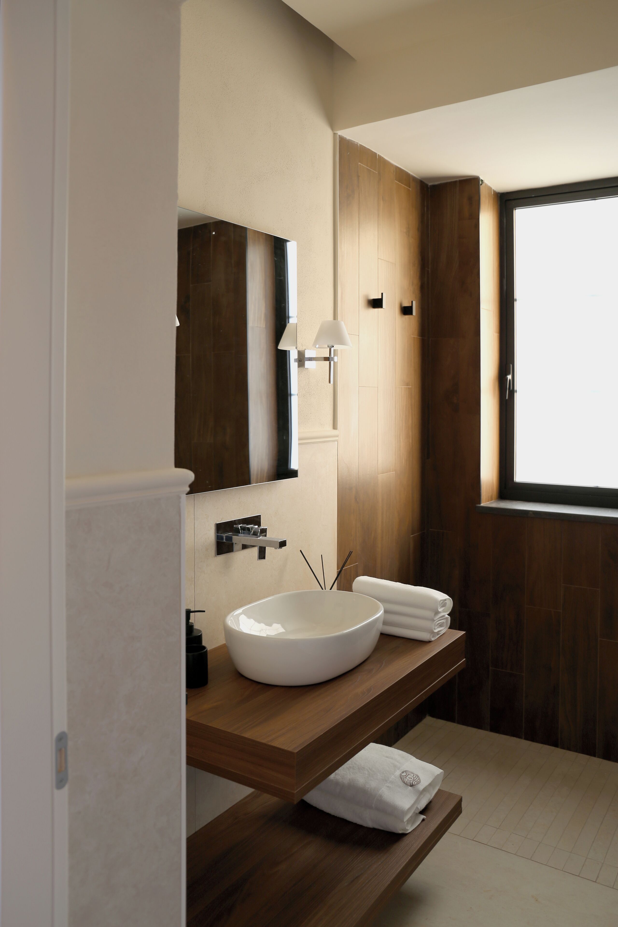 A modern bathroom with a large mirror, a white sink, wooden vanity, and a window providing natural light.