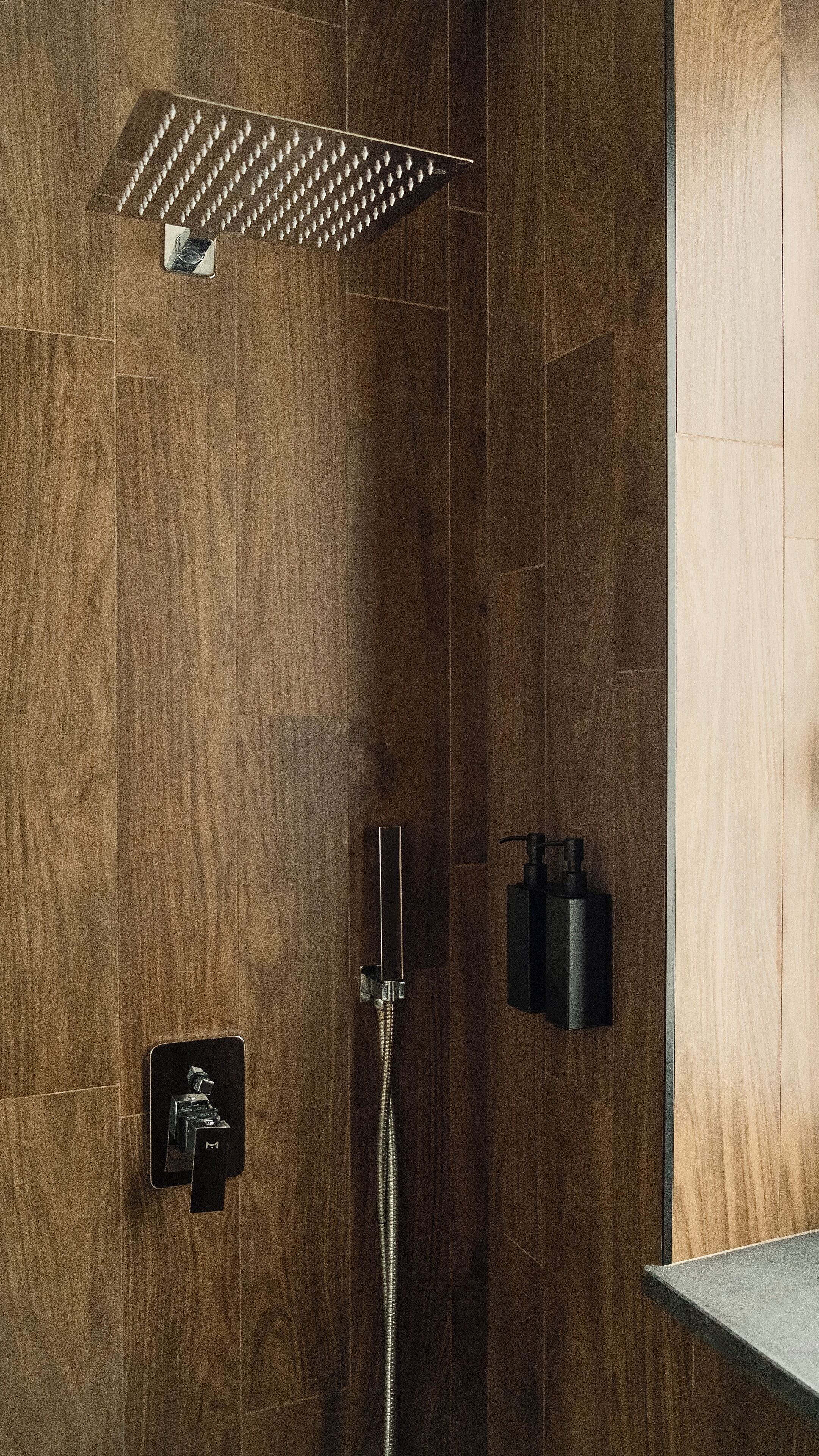 A modern shower with a rain showerhead, a handheld showerhead, and a wall-mounted soap dispenser.