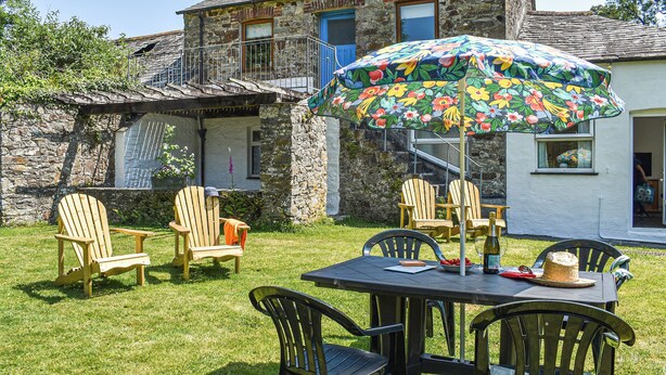Et stenhus med balkon, udendørs siddeplads med stole og et bord, og en farverig parasol.